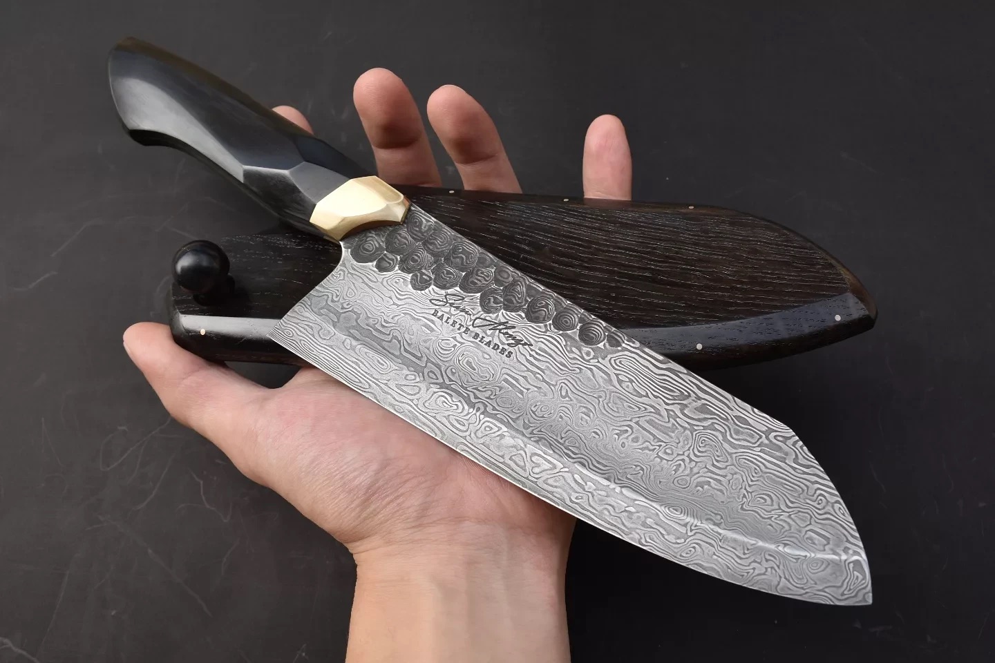 A one-of-one Balete Blades damascus santoku held in the hand beside its wooden saya